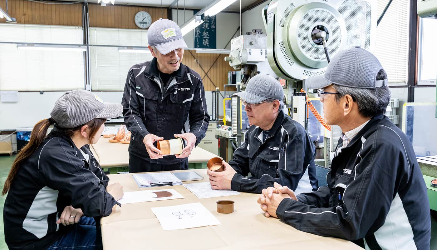 工場内で作業する男女のチームが、それぞれ工具や機器を扱ったり、資料を確認したりしている様子。